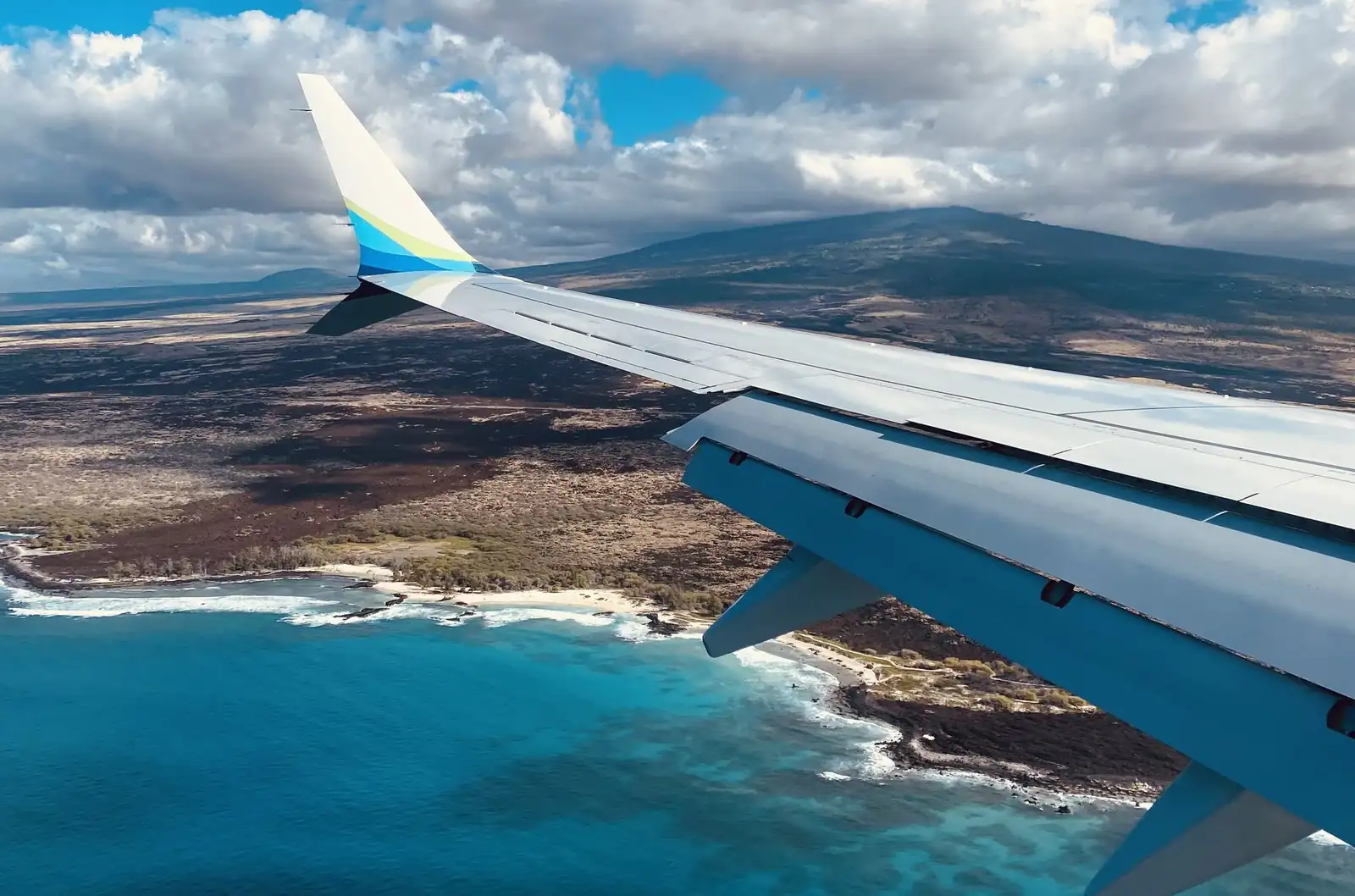 View from a plane window over the Kona coast, with lava fields, turquoise water and Hualālai under the clouds